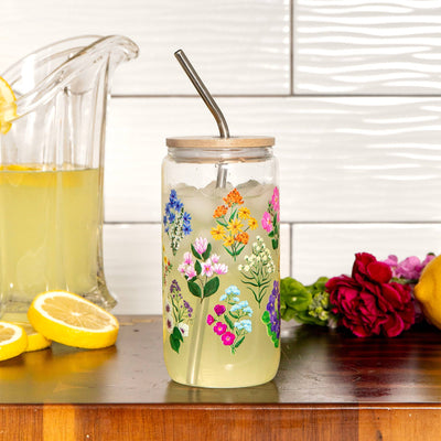 Glass jar with floral design, straw, and wooden lid on a wooden surface with lemons and flowers.
