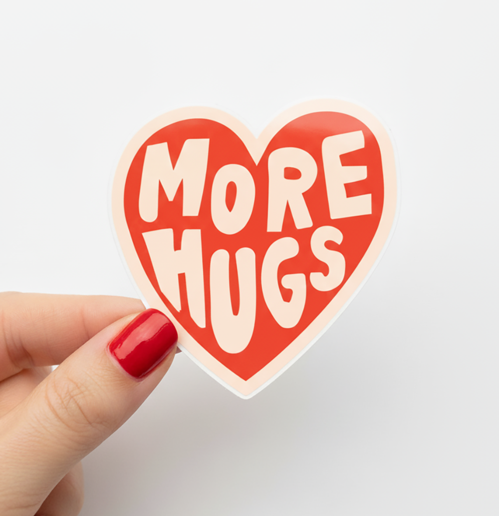 Heart-shaped sticker with 'More Hugs' text held by a hand with red nail polish on a light gray background