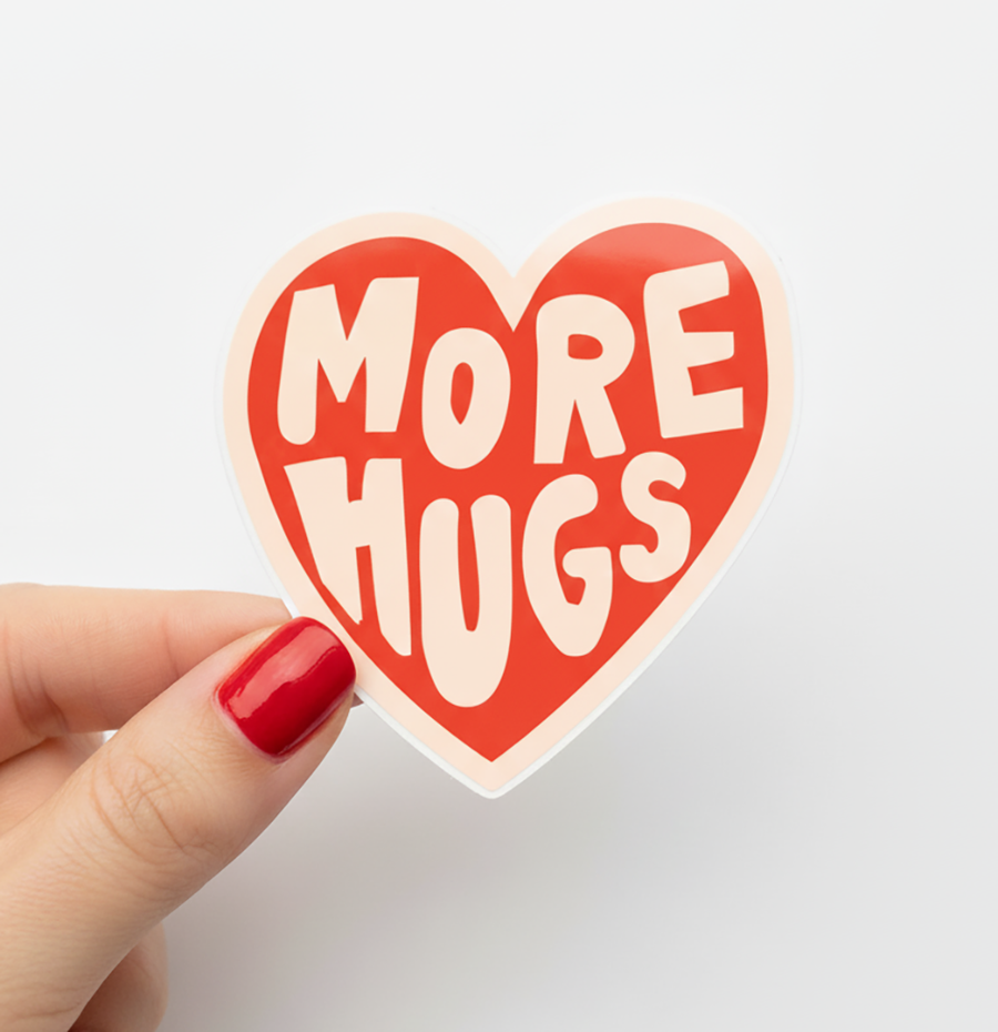 Heart-shaped sticker with 'More Hugs' text held by a hand with red nail polish on a light gray background