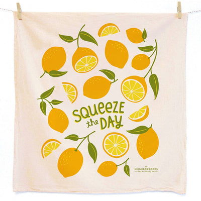 A square beige kitchen towel hanging from two wooden clips against a white background. The towel features a colorful print of lemons and lemon slices with green leaves, surrounding the text 'Squeeze the DAY' in green.