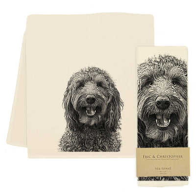 An off-white tea towel with a detailed black-and-white sketch of a dog's face, shown folded with another towel behind it and a matching paper band label on a white background.
