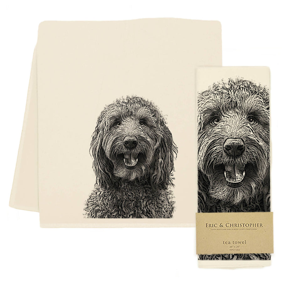 An off-white tea towel with a detailed black-and-white sketch of a dog's face, shown folded with another towel behind it and a matching paper band label on a white background.