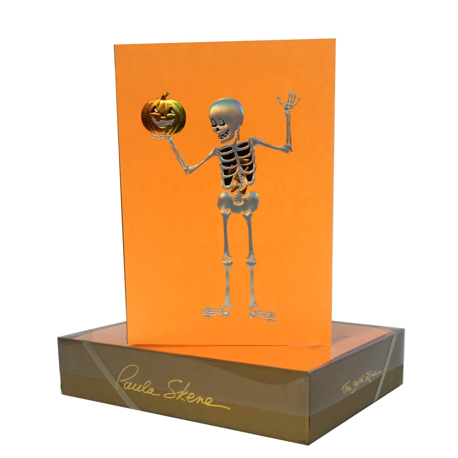 An orange Halloween greeting card featuring a silver skeleton holding a jack-o'-lantern, displayed standing upright in a clear plastic box with a brown base. The box has gold foil script reading 'Paula Skene' and 'The Art of Halloween'.